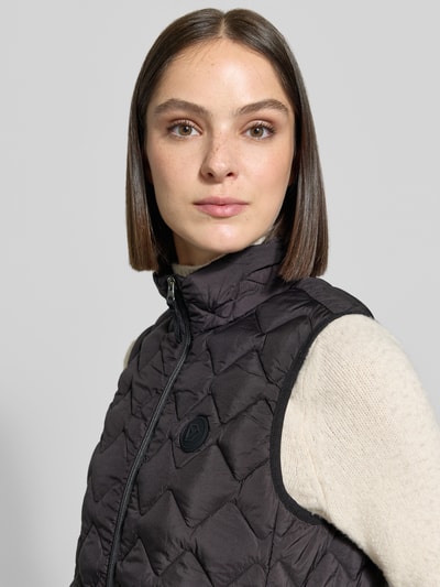 Frauenmodel trägt Regular Fit Weste in Stepp-Optik Modell 'Evy' von Didriksons, schwarz, Stehkragen, Logo, Nahaufnahme Oberkörper. Damen Weste, Größe 36: Rückenlänge 63 cm, Schulterbreite 37 cm, Brustweite 46 cm.