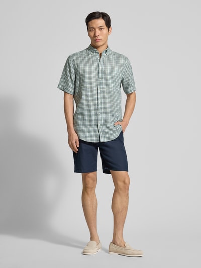 Männermodel trägt Regular Fit vrijetijdsoverhemd uit zuiver linnen van Christian Berg Men met button-downkraag, knoopsluiting, borstzak, groene en blauwe ruit, navy shorts, Ganzkörperansicht van voren. Ruglengte maat M 81 cm, schouderbreedte 54 cm, mouwlengte 28 cm.
