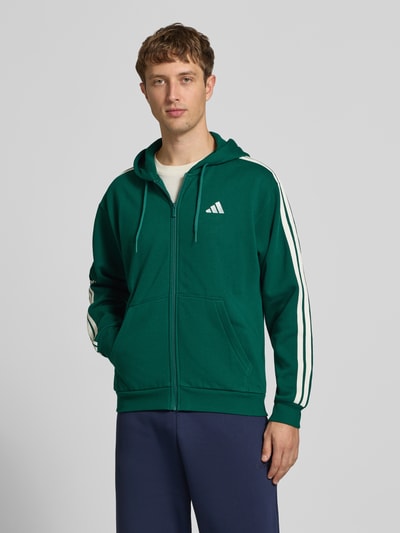 Männermodel trägt Regular Fit Sweatjacke aus Baumwoll-Mix von adidas Sportswear in Grün mit weißen Streifen, Ganzkörperansicht von vorne.