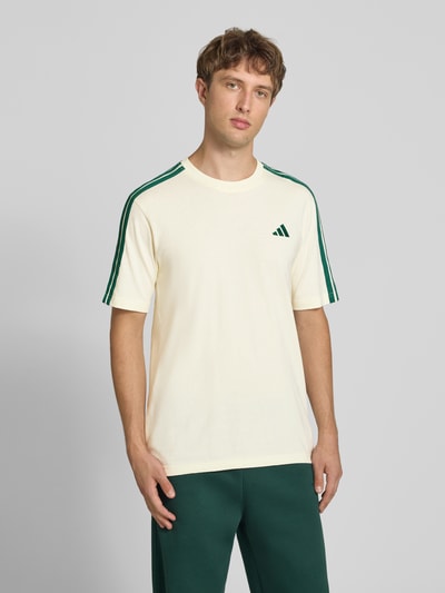 Männermodel trägt Regular Fit T-Shirt aus reiner Baumwolle Modell 'M 3S SJ LC T' von adidas Sportswear in Creme mit grünen Streifen, Ganzkörperansicht von vorne.
