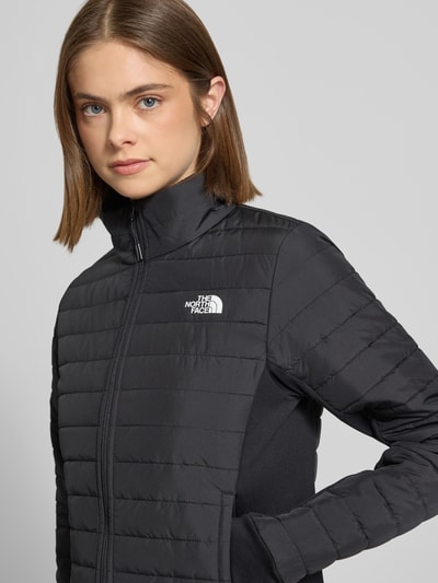 Frauenmodel trägt Steppjacke mit Logo Modell 'CANYONLANDS' von The North Face in Schwarz, glatte Oberfläche, Nahaufnahme Oberkörper. Slim Fit, Stehkragen, Reißverschluss und Reißverschlusstaschen sichtbar, Kontext: Rückenlänge 61 cm, Schulterbreite 36 cm, Brustweite 34 cm, Ärmellänge 70 cm.