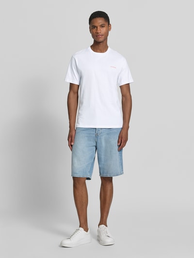 Männermodel trägt T-Shirt mit rückseitigem Motiv-Print von Sundek aus reiner Baumwolle, Regular-Fit, Rundhalsausschnitt, Logo-Print, Rückenlänge bei Größe M 67 cm, weiß, kombiniert mit hellblauer Jeansshorts und weißen Sneakers, Ganzkörperansicht von vorne.