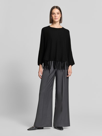 Frauenmodel trägt Relaxed Fit Pullover aus Woll-Mix mit Kaschmir-Anteil von HERZENS, schwarze Strick-Optik mit Fransensaum, Rundhalsausschnitt, kombiniert mit grauer, weiter Stoffhose in neutralen Tönen, Ganzkörperansicht von vorne.