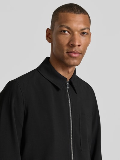 Männermodel trägt Overshirt mit Brusttasche Modell 'JERSON' von Drykorn, schwarzer Material-Mix mit Viskose-Anteil, Modern Fit, mit Kentkragen und Reißverschluss, Nahaufnahme Oberkörper. Rückenlänge bei Größe M: 69 cm.