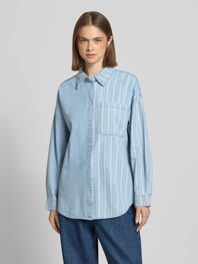 Modelka nosi Oversized Jeansbluse mit Brusttasche Modell 'KATRI' od Noisy May, jasnoniebieska bawełna, oversize, kołnierzyk koszulowy, listwa guzikowa, kieszeń na piersi, obniżone ramiona, mankiety sportowe, zaokrąglony dół, długość tyłu w rozmiarze S 72 cm, długość rękawa w rozmiarze S 50 cm, pełny widok od przodu