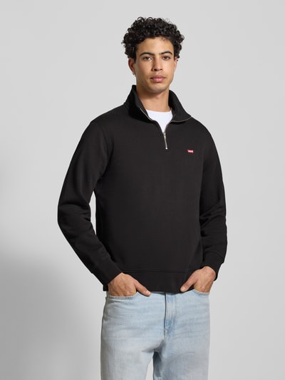 Męski model nosi Sweatshirt mit Label-Detail od Levi's®, czarny kolor, z krótkim suwakiem i stójką, regular fit, widok z przodu na całe ciało.
