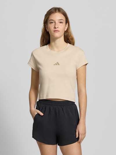 Modelka kobieca ma na sobie Cropped T-Shirt mit Label-Print od adidas Sportswear w beżu, z bawełny, krój slim fit, okrągły ściągacz, nadruk z przodu, długość tyłu w rozmiarze S: 45 cm, widok całej sylwetki z przodu.