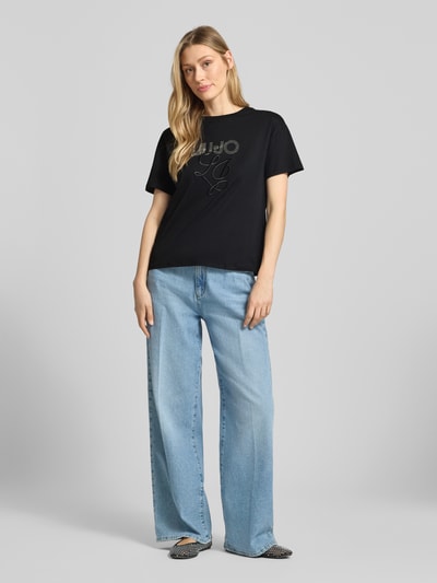 Modelka nosi t-shirt z wyhaftowanym logo i obszyciem ozdobnymi kamieniami od Liu Jo White z bawełny, krój regular fit, okrągły dekolt, długość tyłu w rozmiarze S: 56 cm, widok całej sylwetki z przodu.