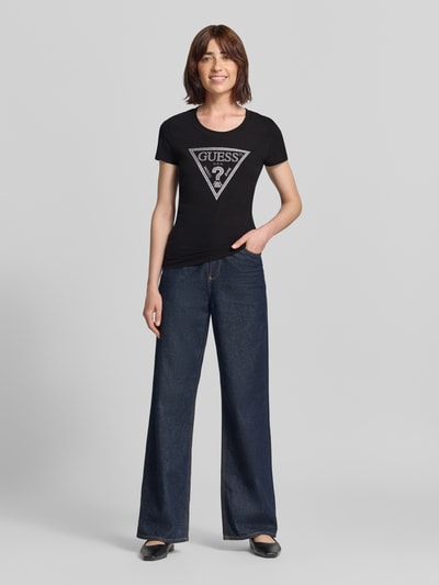 Frauenmodel trägt T-Shirt mit Ziersteinbesatz und Logo von GUESS, Schwarz, Slim Fit aus Baumwoll-Elasthan-Mix, Ganzkörperansicht von vorne.