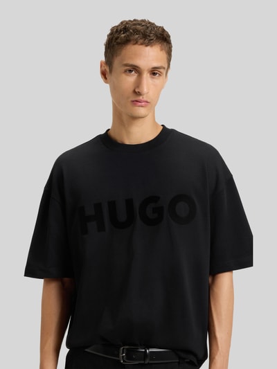 Männermodel trägt Oversized T-Shirt aus reiner Baumwolle Modell 'DINKEE' von HUGO in Schwarz, unifarben mit Label Print, Nahaufnahme Oberkörper.