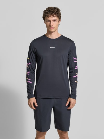 Männermodel trägt Funktionsshirt mit Logo Modell 'Kout' von Salomon, Regular Fit, Longsleeve, dunkel, dezente Farbakzente, Ganzkörperansicht von vorne