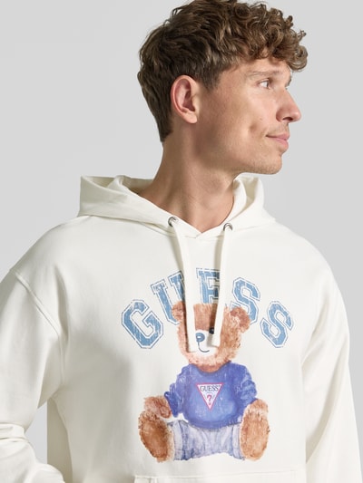 Männermodel trägt Hoodie mit Label-Print und Känguru-Tasche von GUESS, Weiß mit blau-braunem Print, Baumwoll-Mix, Nahaufnahme Oberkörper.