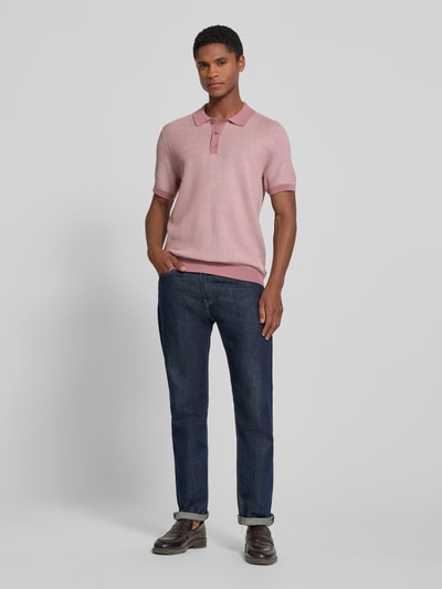 Męskamodel nosi koszulkę polo Slim Fit Poloshirt mit Strukturmuster model 'Brianoro' von JOOP! COLLECTION z bawełny, krój slim fit, fakturowany wzór, listwa guzikowa, detal z logo, długość tyłu w rozmiarze M: 70 cm, ciemnoniebieskie spodnie, kolor różowy, widok całej sylwetki z przodu.