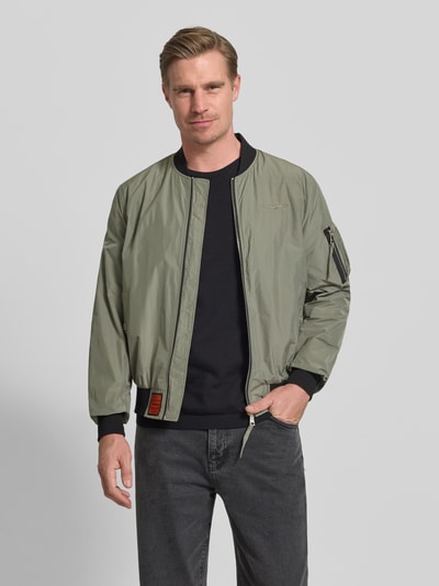 Männermodel trägt Bomberjacke mit Label-Patch und Eingrifftaschen von Original Bombers in Olivgrün mit Reißverschluss und gerippten Abschlüssen, Ganzkörperansicht von vorne. Gerader Schnitt, Reißverschlusstasche am Ärmel, Druckknopf-Taschen, Maße: Rückenlänge 67 cm, Schulterbreite 43 cm, Brustweite 53 cm, Ärmellänge 68 cm.