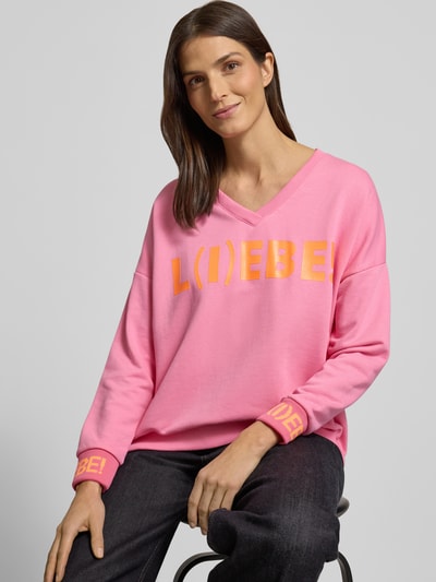 Frauenmodel trägt Oversized Sweatshirt mit Statement-Print von miss goodlife in Pink mit gelbem Schriftzug, reine Baumwolle, gerippte Abschlüsse, V-Ausschnitt, Ganzkörperansicht von vorne.