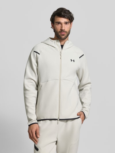Männermodel trägt Regular Fit Jacke mit Kapuze Modell 'Unstoppable' von Under Armour in Creme mit Logo-Print, Zweiwege-Reißverschluss, Elastischer-Material-Mix, geteilte Känguru-Tasche, elastische Abschlüsse, Rückenlänge 75 cm, Ärmellänge 57 cm, Nahaufnahme Oberkörper.