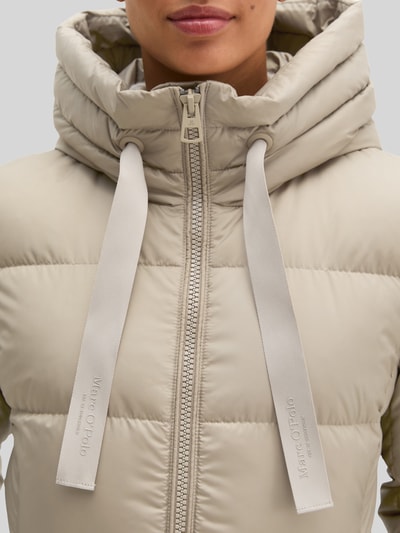 Kobieta nosi Regular Fit Daunenmantel mit Label-Print od Marc O'Polo, jasnobeżowy, matowa tekstura, z widocznym nadrukiem z logo, zbliżenie na górną część płaszcza, kontekst: damski płaszcz puchowy od Marc O'Polo, zbliżenie na górę