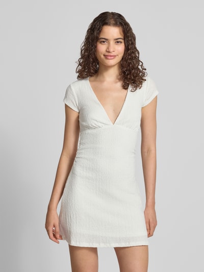Frauenmodel trägt Minikleid mit V-Ausschnitt von Gina Tricot in Weiß, Baumwoll-Mix mit Stretch, Strukturmuster, Ganzkörperansicht von vorne.