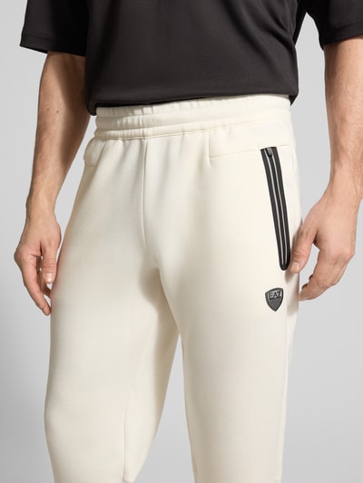 Männermodel trägt cremefarbene Sweatpants mit Viskose-Anteil Modell 'TRAIN PREMIUM SHIELD' von EA7 Emporio Armani, Nahaufnahme Oberkörper. Elastischer Bund, Reißverschlusstaschen und gerippte Abschlüsse, gerade Passform, detailreiche Materialstruktur.
