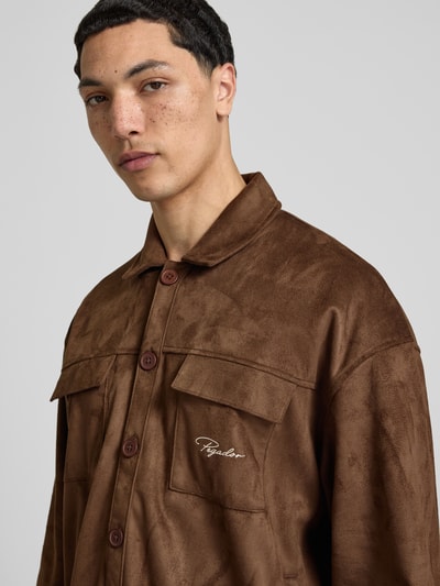 Mężczyzna model nosi Relaxed Fit Hemdjacke mit Brusttaschen Modell 'Madikeri' von Pegador w kolorze brązowym, faktura imitująca skórę, naszyte kieszenie na piersi, kent kołnierzyk, brązowe guziki, delikatny haft z logo firmy, długość tyłu w rozmiarze M: 69 cm, zbliżenie górnej części ciała z przodu.