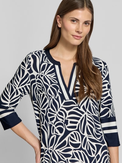 Modelka kobieta ma na sobie knielanges Tunikakleid mit V-Ausschnitt und 3/4-Arm von Christian Berg Woman Selection, wzór allover, mieszanka materiałów z domieszką stretchu, prosty krój, długość do kolan, szerokość w rozmiarze 36: 41 cm, długość tyłu: 92 cm, długość rękawa: 42 cm, ciemnogranatowe i białe liście, widoczne tekstury, zbliżenie na górną część ciała, widok z przodu.