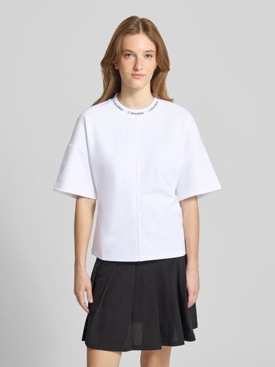 Frauenmodel trägt weißes Comfort Fit T-Shirt mit Label-Schriftzug von Calvin Klein Jeans, kombiniert mit schwarzem Rock, Ganzkörperansicht von vorne.