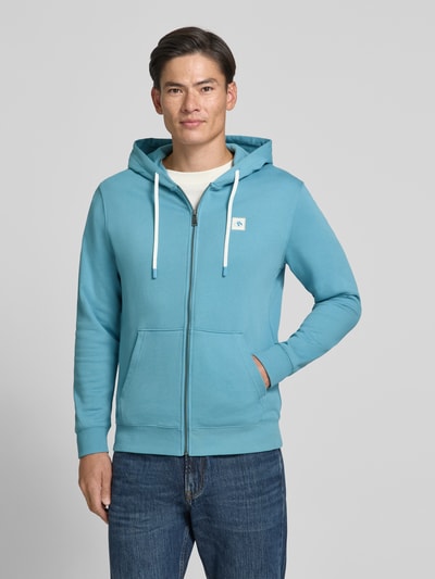 Männermodel trägt Sweatjacke mit Logo-Patch und Känguru-Tasche von Scotch & Soda in Hellblau mit Kapuze und Reißverschluss, Ganzkörperansicht von vorne. Baumwollmaterial, gerader Schnitt, gerippte Abschlüsse, Kontext: Herren Sweatjacke von Scotch & Soda, Rückenlänge 70 cm bei Größe M, Ärmellänge 68 cm bei Größe M.