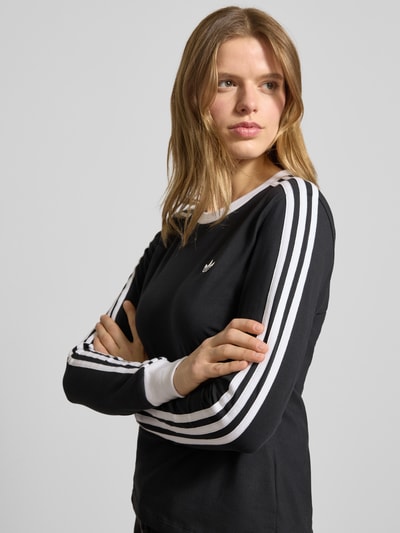 Kobiecy model nosi Regular Fit Langarmshirt aus Baumwoll-Mix von adidas Originals, efekt: jednolity kolor, paski w kontrastowym kolorze, wyhaftowane logo, okrągły dekolt, długi rękaw, ściągacze, mieszanka bawełny, zbliżenie na górną część ciała z przodu.