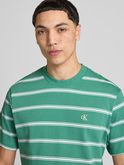 Männermodel trägt T-Shirt mit Logo-Stitching von Calvin Klein Jeans in Grün mit weißen Streifen aus Reine Baumwolle, Gerader-Schnitt, Rundhalsausschnitt, Rückenlänge bei Größe M: 69 cm, Nahaufnahme Oberkörper.