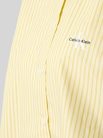 Frauenmodel trägt Regular Fit Pyjama-Oberteil aus reiner Baumwolle von Calvin Klein, gelb gestreift, Popeline, Knopfleiste, Nahaufnahme Oberkörper.