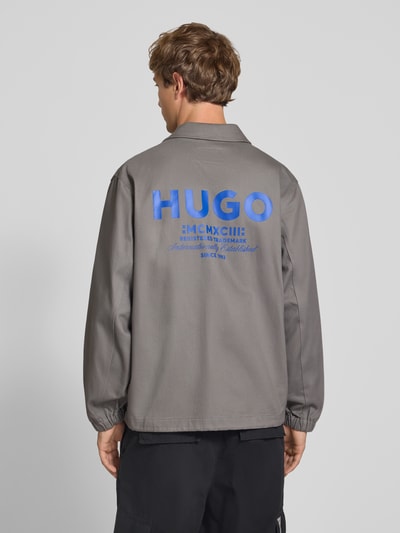 Männermodel trägt Relaxed Fit Blouson aus reiner Baumwolle Modell 'Biyo2631' von HUGO, grauer Stoff mit blauem Logo, Ganzkörperansicht Rückansicht. Herren Bomberjacke / Blouson von HUGO Blue