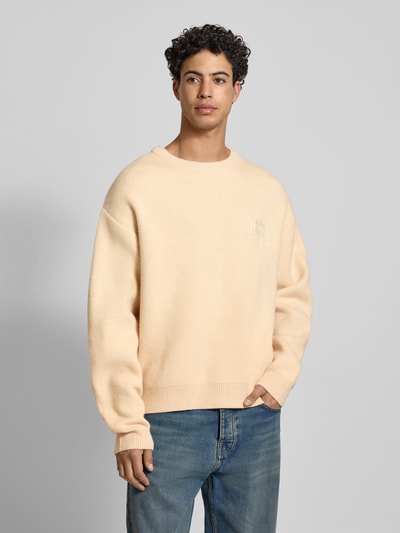 Männermodel trägt Strickpullover mit gerippten Abschlüssen von ANOTHER COTTON LAB, Material-Mix, lockerer Schnitt, Rundhalsausschnitt, Label-Stitching, beige Farbe, mittlere Struktur, Ganzkörperansicht von vorne.