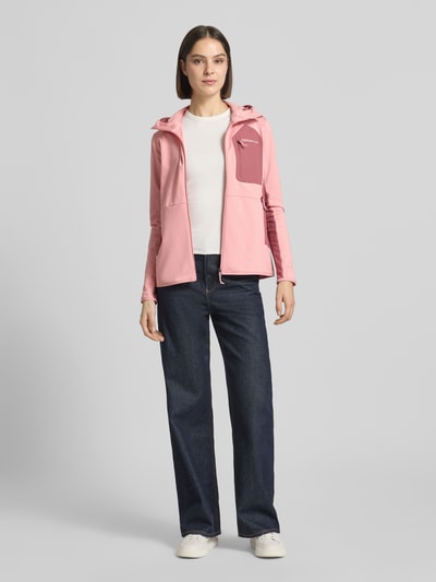 Frauenmodel trägt Jacke mit Logo-Stitching von PEAK PERFORMANCE in Rosa, Elastischer-Material-Mix, Körpernahe Passform, Kapuze, Reißverschluss, Brusttasche mit Reißverschluss, Ganzkörperansicht von vorne.