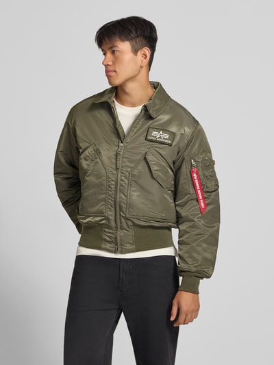 Männermodel trägt grüne Bomberjacke mit seitlichen Pattentaschen und Label-Patch von Alpha Industries, glatte Oberfläche, Ganzkörperansicht von vorne. Regular Fit, Kentkragen, Reißverschluss, Reißverschlusstasche am Ärmel mit Label-Detail, gerippte Abschlüsse, Rückenlänge 62 cm, Schulterbreite 50 cm, Brustweite 61 cm, Ärmellänge 67 cm.