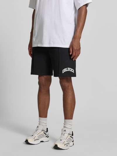 Männermodel trägt Regular Fit Shorts mit Tunnelzug von COLUCCI aus reiner Baumwolle, Schwarz mit Label-Stitching, Nahaufnahme Unterkörper.