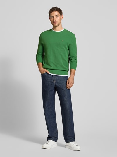 Męski model nosi zielony Regular Fit Pullover aus reiner Baumwolle von Tommy Hilfiger, ciemnoniebieskie jeansy, białe buty, Ganzkörperansicht von vorne.