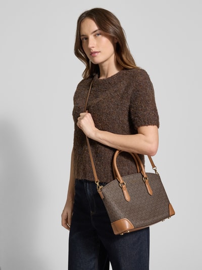 Frauenmodel trägt Handtasche mit Logo-Applikation Modell 'Murphey' von MICHAEL Michael Kors, brauner Allover-Logo-Print, Schulterhenkel, Ganzkörperansicht von vorne.