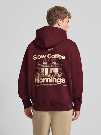 Männermodel trägt Regular Fit Hoodie aus Baumwoll-Mix von PEGADOR in Dunkelrot mit beigefarbenem Print, gerippte Abschlüsse, Kängurutasche, Strass-Logo, Rückenansicht, Ganzkörperansicht Rückansicht.