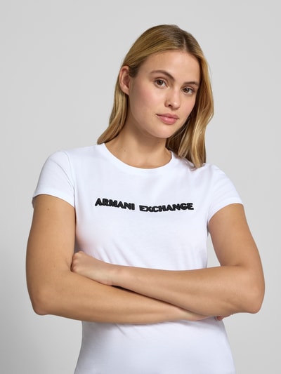 Kobiece modelka nosi Slim Fit T-Shirt aus reiner Baumwolle von Armani Exchange, biały kolor, detal z logo, okrągły dekolt, damski t-shirt, długość tyłu w rozmiarze S: 59 cm, krój slim fit, bawełna, detal z logo, okrągły dekolt, zbliżenie na górną część ciała.