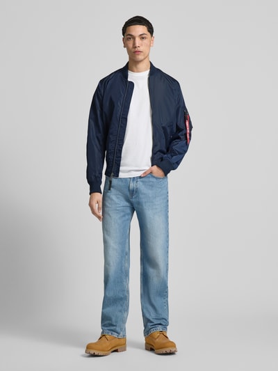 Męski model trägt Regular Fit Jacke mit Stehkragen von Alpha Industries w kolorze granatowym, wpuszczane kieszenie, kieszeń na rękawie, zamek błyskawiczny, elastyczne wykończenia, długość tyłu w rozmiarze M 66 cm, szerokość ramion w rozmiarze M 48 cm, szerokość klatki piersiowej w rozmiarze M 54 cm, długość rękawów w rozmiarze M 68 cm, Ganzkörperansicht von vorne.