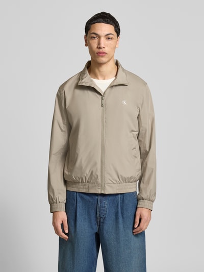 Männermodel trägt Trainingsjacke mit Logo und Stehkragen von Calvin Klein Jeans, regular fit, Reißverschluss, Eingrifftaschen, beige, Ganzkörperansicht von vorne