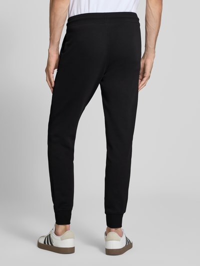 Mężczyzna nosi spodnie dresowe Sweatpants mit Label-Print Modell 'GORDON' od JACK & JONES, czarny kolor, wpuszczane kieszenie, elastyczny pas z tunelem, luźny krój, długość wewnętrzna nogawki w rozmiarze M: 77 cm, widok całej sylwetki z tyłu.