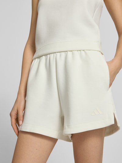 Frauenmodel trägt Shorts mit Label-Print von adidas Sportswear in Cremeweiß, lockerer Schnitt, Material-Mix, Nahaufnahme Oberkörper.