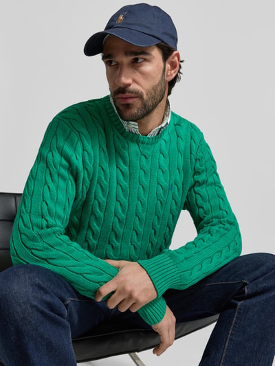 Männermodel trägt Regular Fit Strickpullover aus reiner Baumwolle von Polo Ralph Lauren, Optik: Unifarben, Strickoptik, Zopfmuster, Logo-Detail, Rundhals, Langarm, gerippte Abschlüsse, Materialverarbeitung: Strick, Farbe: Grün; Ganzkörperansicht von vorne.
