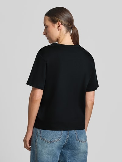 Kobieta nosi Damski t-shirt Regular Fit T-Shirt aus reiner Baumwolle Model 'Melea' od MSCH Copenhagen, czarny kolor, krój regular fit, nadruk z logo, okrągły dekolt, bawełna, długość tyłu w rozmiarze XS/S: 57 cm, widok całej sylwetki od tyłu.