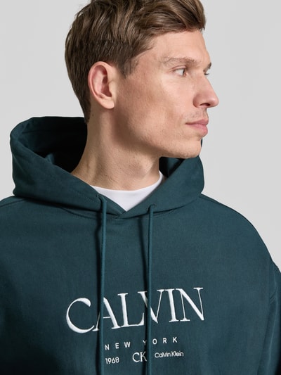 Männermodel trägt Hoodie mit Label-Print und Känguru-Tasche van CK Calvin Klein in donkergroen, mix van katoen en elastaan, relaxed fit, labelprint en kangoeroezak, ruglengte bij maat M 69 cm, mouwlengte bij maat M 60 cm, Nahaufnahme Oberkörper.