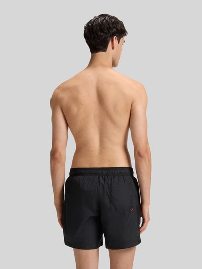 Männermodel trägt schwarze Badeshorts mit Label Print Modell 'HAITI' von HUGO, elastischer Bund, Gesäßtasche, Ganzkörperansicht Rückansicht.