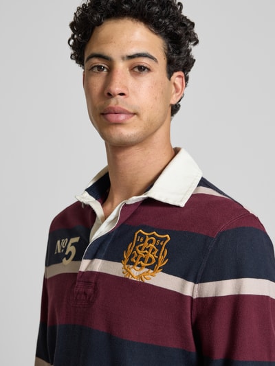 Männermodel trägt Regular Fit Rugby Shirt mit Logo- und Motiv-Stitching von Superdry in Bordeaux, Navy und Beige, Nahaufnahme Oberkörper. Reine Baumwolle, Polokragen, kurze Knopfleiste, Label-Patch sichtbar.