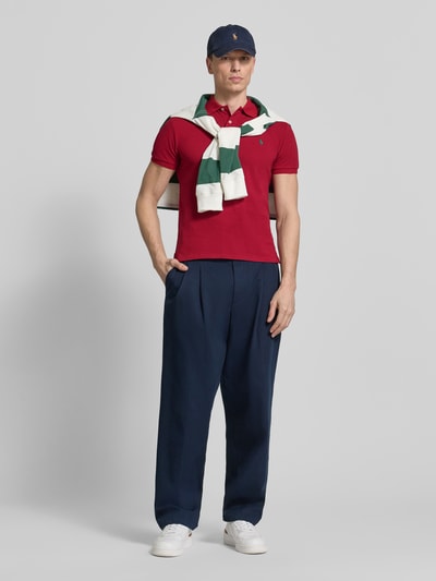 Mannenmodel draagt 'Slim Fit Poloshirt aus reiner Baumwolle' van Polo Ralph Lauren in effen rood, piqué katoen, logo-stiksel, polokraag, korte mouw, zijsplit, twee knopen, slim fit ontwerp met casual navy broek en witte sneakers. Ganzkörperansicht van voren.