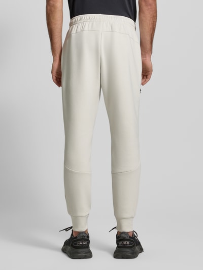 Männermodel trägt Regular Fit Sweatpants mit Logo-Print Modell 'Unstoppable' von Under Armour in Beige, elastischer Material-Mix mit Baumwoll-Anteil, Beintasche mit Reißverschluss, elastischer Bund und Beinabschlüsse, Innenbeinlänge 73 cm in Größe M, Ganzkörperansicht Rückansicht.